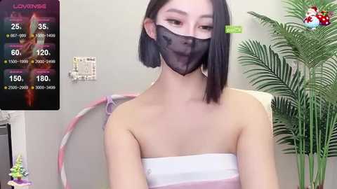 Media: Video of an East Asian woman with straight black hair and a white strapless dress, wearing a black face mask. Background features a green plant, pink hula hoop, and a digital screen displaying health stats.