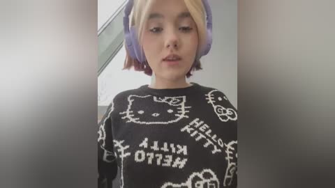 Media: A video of a young woman with light skin, short blonde hair, and wearing purple headphones, a black sweater featuring Hello Kitty designs, and a neutral expression.