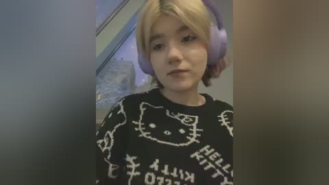 A young woman with light skin and short, blonde hair wears large purple headphones and a black \"Hello Kitty\" sweater indoors, with blurred background.