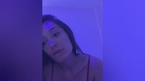 Media: Video of a young woman with shoulder-length dark hair, wearing a red spaghetti strap top, standing against a purple-lit background, looking introspective.