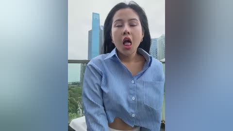 Media: A video of an Asian woman with long black hair, wearing a blue striped button-up shirt, standing on a balcony, mouth open in a surprised expression, with a city skyline in the background.