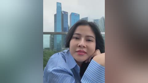 Media: A video of an East Asian woman with straight black hair and fair skin, wearing a blue striped shirt, leaning on a balcony overlooking modern skyscrapers and greenery under a cloudy sky.