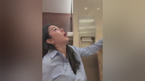 Media: Video of an Asian woman with long black hair, wearing a blue and white striped button-up shirt, leaning against a beige stall in a public restroom.