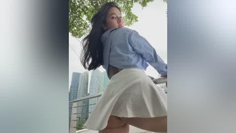 Media: Video of a young woman with long black hair, wearing a blue plaid shirt and white mini skirt, posing seductively outdoors by a building with green trees and modern skyscrapers in the background.