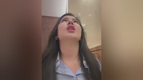 Media: Video of a woman with long dark hair and light skin, wearing a striped blue shirt, leaning against a beige wall, mouth slightly open, looking upwards, in an indoor setting.