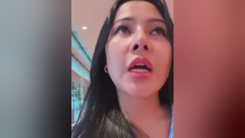 Media: A close-up video of a young woman with long black hair, light skin, and red lipstick, captured indoors against a blurred background, appearing to be in a reflective mood.