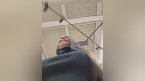 Media: Video of a smiling woman with long hair, wearing a dark sweater, standing in a bright, industrial room with white walls and exposed metal beams.