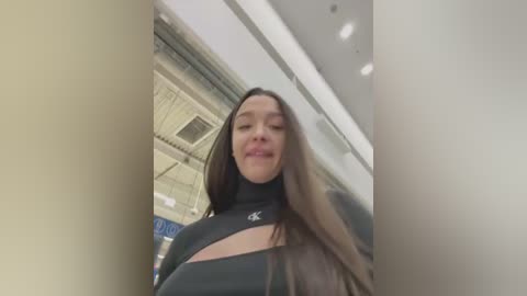 Media: A video of a young woman with long brown hair, wearing a black top, smiling, taken from a low angle in a modern, brightly lit indoor setting.