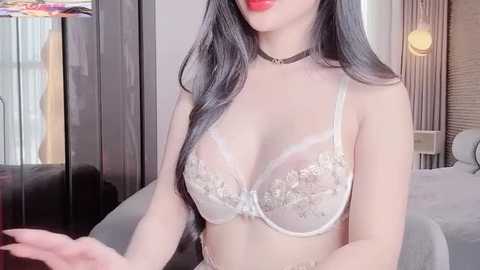 Video of an Asian woman with long black hair, wearing a sheer white lace bra revealing medium-sized breasts, a black choker, and red lipstick, in a modern, softly lit bedroom.