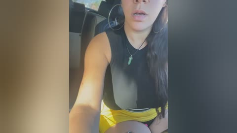 Media: Video of a young woman with medium skin tone, long dark hair, wearing a black sleeveless top and yellow skirt, seated in a car, visible in the background.