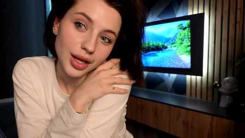 Media: Video of a young woman with short brown hair, light skin, and blue eyes, wearing a white sweater, leaning forward with her chin on her hand, in a modern living room with a TV displaying a nature scene.