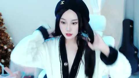 Media: Video of an East Asian woman with fair skin and long black hair, wearing a black beret and a white, black-trimmed coat, adjusting her hair. Background includes a white bed with a decorative pillow and a partially visible Christmas tree.