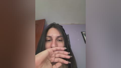 Media: Video of a young woman with long, dark hair, fair skin, and almond-shaped eyes, partially obscured by her hand, in a room with lavender walls and a framed picture.