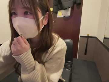 Media: Video of an East Asian woman with long brown hair, wearing a white face mask, holding a tissue to her mouth, sitting in a medical examination room with beige walls and black furniture.