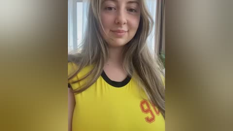 Video of a young woman with long, straight blonde hair, wearing a yellow soccer jersey with black collar and \"99\" in red on the chest. She has a slight smile, fair skin, and is indoors with a blurred background.