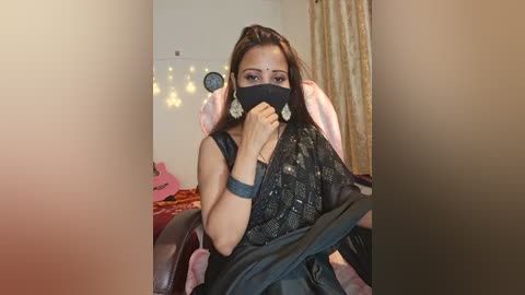 Media: A video of a woman in a black mask and saree, sitting on a pink gaming chair, with a background of string lights and a pink toy.