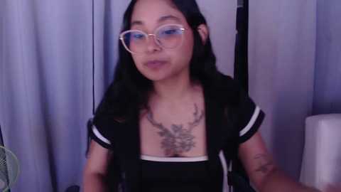 Media: Video of an East Asian woman with long black hair, wearing clear glasses, black top, and visible tattoos on her chest, standing in front of light purple curtains.