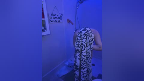 Media: A video under blue lighting shows a person with a tattooed backside, wearing patterned pants, standing in a dimly lit bathroom with a showerhead and a motivational sign.