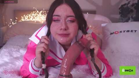 Media: A video depicts a young woman with long, dark hair, wearing a pink outfit, performing oral sex on a large, erect penis, on a white, fluffy bed in a softly lit room.