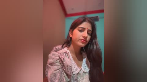 Media: Video of a young South Asian woman with long dark hair, wearing a traditional saree with intricate embroidery, standing in a softly lit room with red and teal accents.