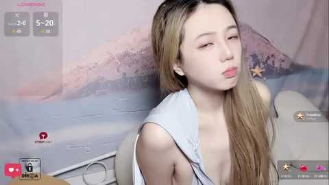 Media: Video of a young Asian woman with long, straight, blonde hair, wearing a sleeveless white top, sitting indoors. The background features a pastel mural of a beach and palm trees.