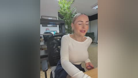 Media: Video of a smiling, blonde woman with fair skin in a white long-sleeve top, sitting at a wooden table in a modern, well-lit kitchen with green plants and a black fridge.