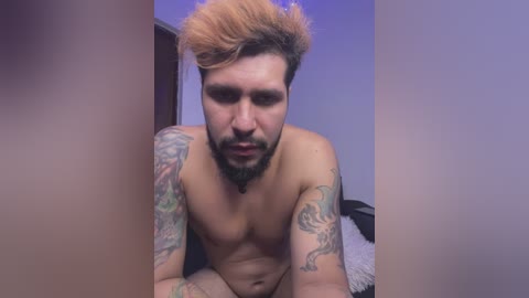 Media: Video of a shirtless, muscular, tattooed man with light skin and short, bleached hair, gazing intensely at the camera. He's leaning forward on a bed with white sheets in a dimly lit room.