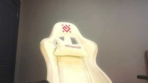 Media: Video of a white gaming chair with a red \"Razer\" logo and a small pillow, placed against a plain beige wall. The chair has a sleek, ergonomic design with adjustable armrests.