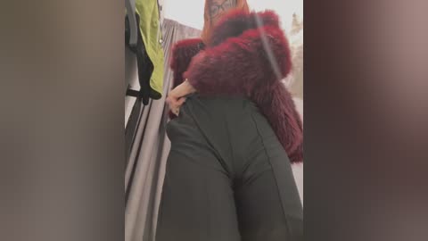 Media: Video of a woman in a red fur coat, green leggings, and a tattooed arm, bending over in a store, partially obscured by a blurry background.