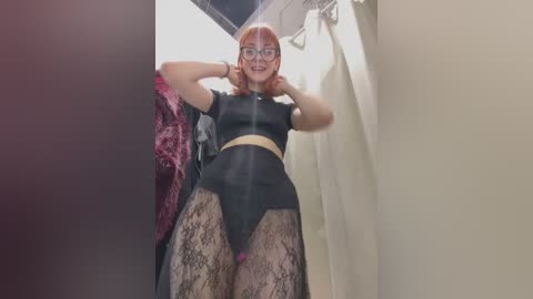 Video of a light-skinned woman with red hair, wearing glasses, black lace lingerie, and high-waisted panties, posing in a narrow, dimly lit hallway.