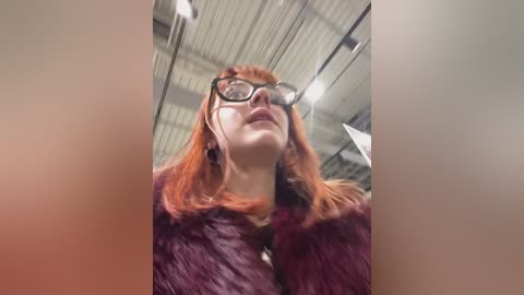 A video of a young woman with red hair, wearing glasses and a maroon fur coat, standing indoors under a metallic ceiling, looking upward.