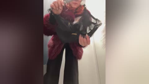 Video of a person in a maroon fur coat, black top, and black pants, holding a black purse, wearing glasses, indoors with a blurred background.