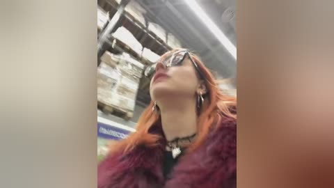 Media: Video of a young woman with long, orange hair, wearing black-framed glasses, a fur coat, and a black necklace, looking upward in a dimly lit, industrial setting with exposed pipes and fluorescent lighting.