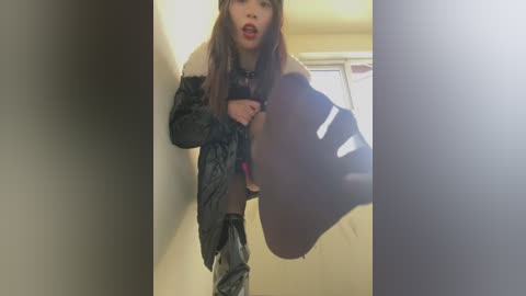 Media: Video of a young woman with long brown hair, wearing a black leather jacket, black thigh-high boots, and a short skirt, leaning against a wall in a dimly lit hallway.