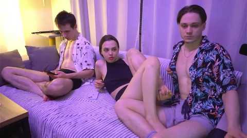 Media: Video of three young people on a striped sofa in a dimly lit room. Two men, one in a floral shirt and the other in a white shirt, are seated. A woman in a black top and black shorts is lying on her side, looking at her phone.