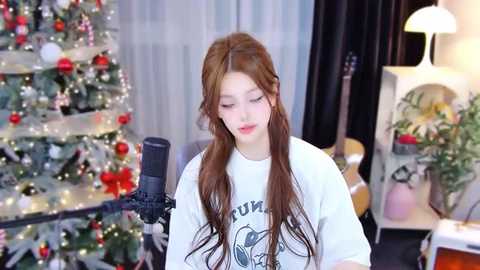 Media: Video of a young woman with long, wavy brown hair, wearing a white t-shirt, singing into a microphone in a cozy, festive room with a decorated Christmas tree and a guitar.