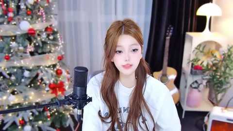 Media: Video of a young Asian woman with long, wavy brown hair, wearing a white graphic t-shirt, singing into a microphone in a brightly lit room with a decorated Christmas tree and guitar in the background.