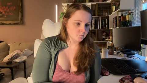 Video of a fair-skinned woman with long brown hair, wearing a pink lace bra and a green cardigan, sitting at a cluttered desk in a cozy, book-filled room.