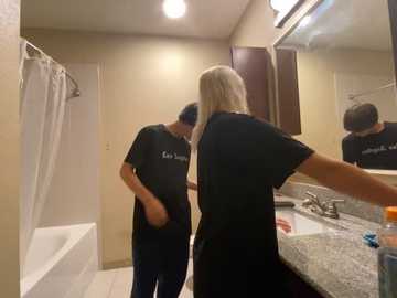 Media: Video of a blonde woman in a black t-shirt and jeans, facing a bathroom mirror, adjusting her hair, while a man in a black shirt and jeans stands beside her.