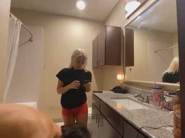 Media: Video of a blonde woman with fair skin in a bathroom, wearing a black t-shirt and red shorts, looking at a smartphone, with a granite countertop, mirror, and toiletries in the background.