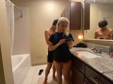 Media: Video of a young blonde woman in a black shirt and red shorts, standing in a modern bathroom, holding a phone, with a shirtless man behind her, reflecting in the mirror.