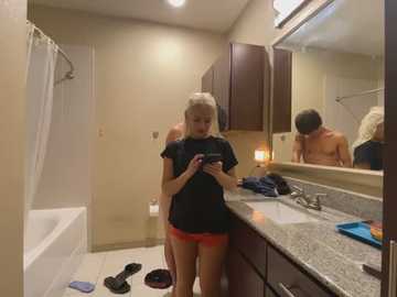 Media: Video of a blonde woman in a bathroom, wearing a black T-shirt and red shorts, texting, surrounded by clutter, including a bathtub, clothes, and toiletries.