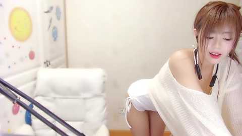 Media: Video of an Asian woman with straight, shoulder-length brown hair, wearing a loose white off-shoulder sweater and black headphones. She stands in a modern, minimalist room with white walls and a white leather couch, with colorful circular stickers on the wall.