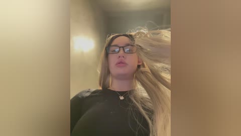 Media: Video of a young woman with long, blonde hair, wearing black-framed glasses, a black top, and a heart pendant necklace. She's in a dimly lit hallway, blowing hair away from her face.