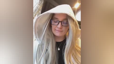 Video of a young woman with long, straight blonde hair, wearing black-framed glasses, a black top, and a white hoodie with hood up, indoors with a blurred background.