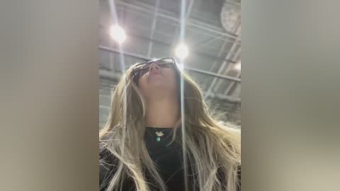 Media: A video of a blonde woman with long hair, wearing sunglasses and a black top, standing in an industrial warehouse with exposed beams and bright lights.