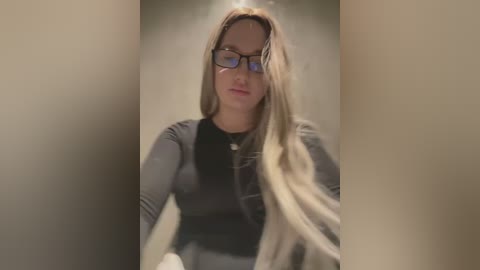 Media: Video of a young Caucasian woman with long blonde hair, wearing black-framed glasses and a black top, seated in a dimly lit, beige-colored room.