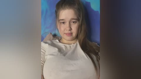 Media: Video of a young Caucasian woman with light skin, straight brown hair, wearing a white top, against a blue background. She has a neutral expression.