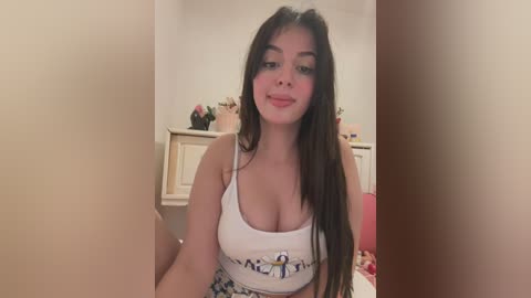 Media: Video of a young woman with long, straight black hair, fair skin, and full lips, wearing a white crop top that accentuates her ample breasts. She sits in a cozy, softly lit bedroom with a pink bedspread in the background.