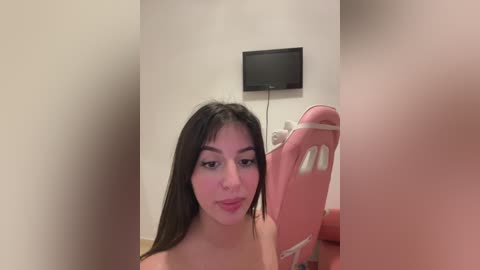 Media: Video of a young woman with long black hair, fair skin, and a slim build, standing in a beige room with a flat-screen TV on the wall and a pink gaming chair.